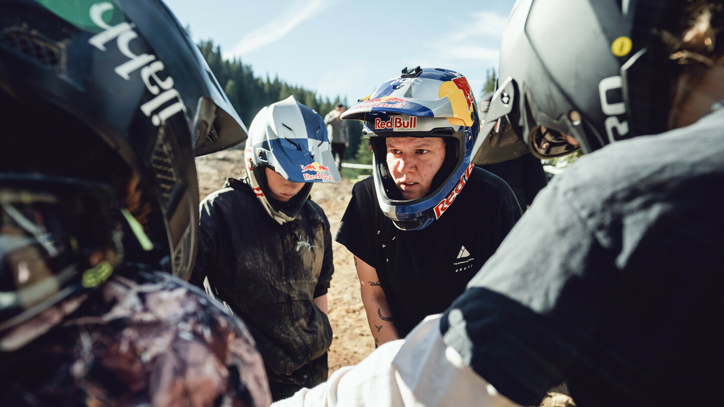 A group of mountain bikers, all wearing helmets, huddled together in discussion. They are wearing various helmets, and are in sporty gear.